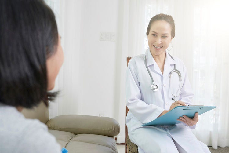 Smiling Asian female doctor in white coat sitting on chair prescribing a medicine for patient while visiting her at home (Smiling Asian female doctor in white coat sitting on chair prescribing a medicine for patient while visiting her at home, ASCII, 