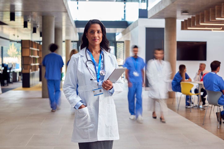 Portrait Of Female Doctor Wearing White Coat With Digital Tablet In Busy Hospital